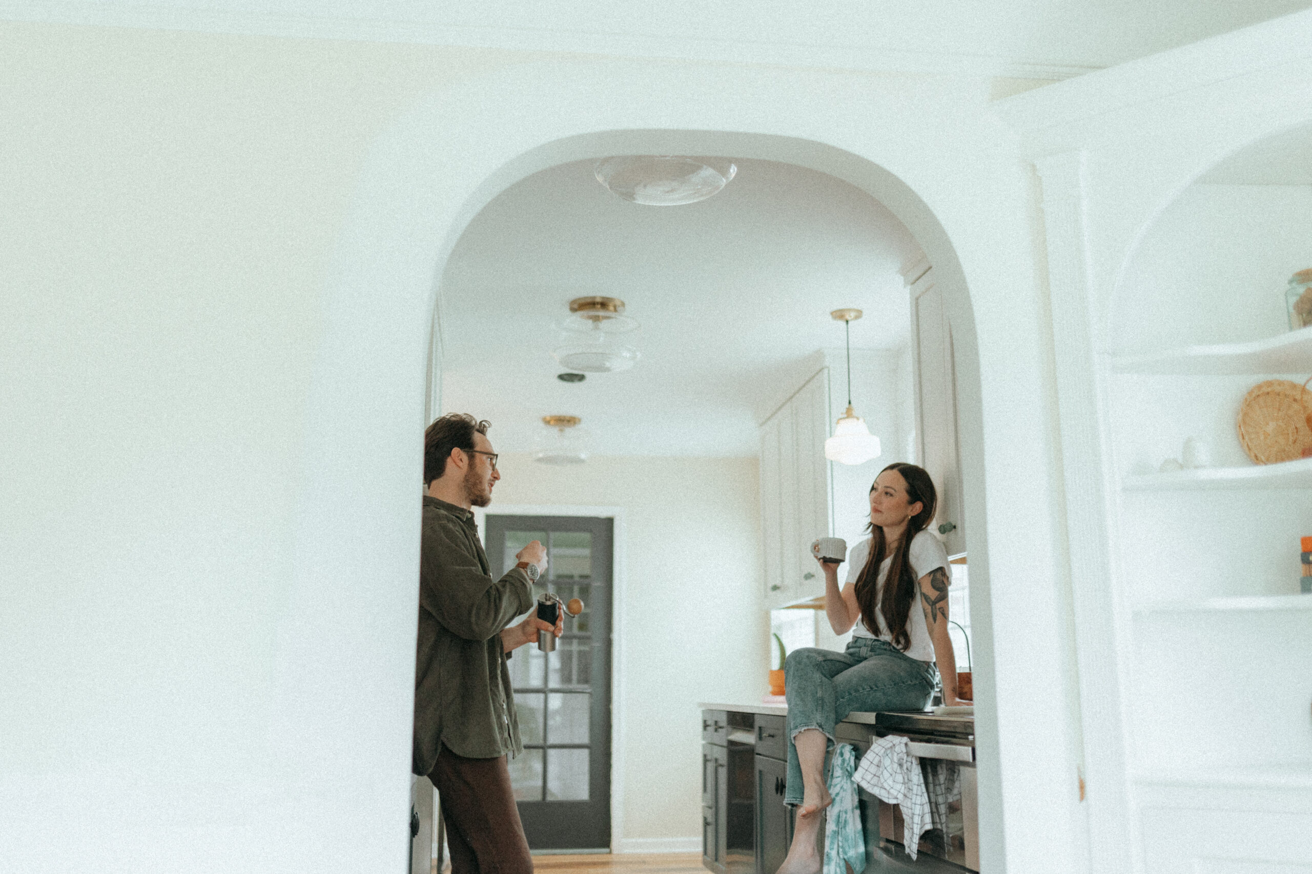 Michigan wedding photographer at home couple engagement session michigan photographer Guy standing making coffee across from girl sitting on kitchen counter drinking coffee